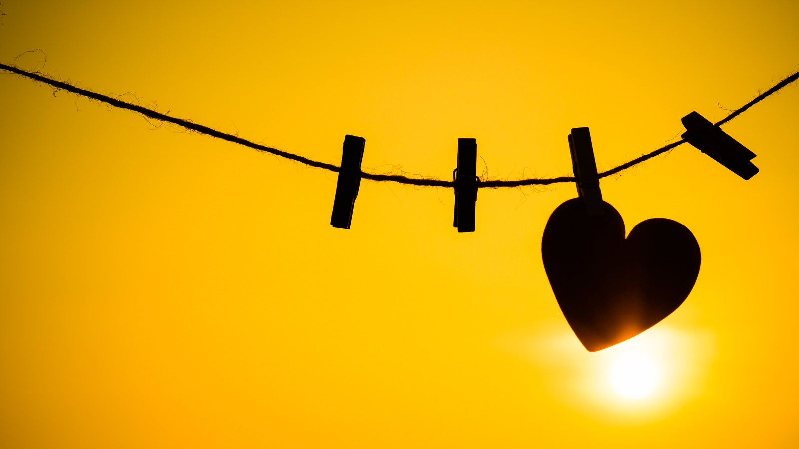 Silhouette of a heart shape being held on a string by a clothing peg. The background shows the sun shining against a yellow sky