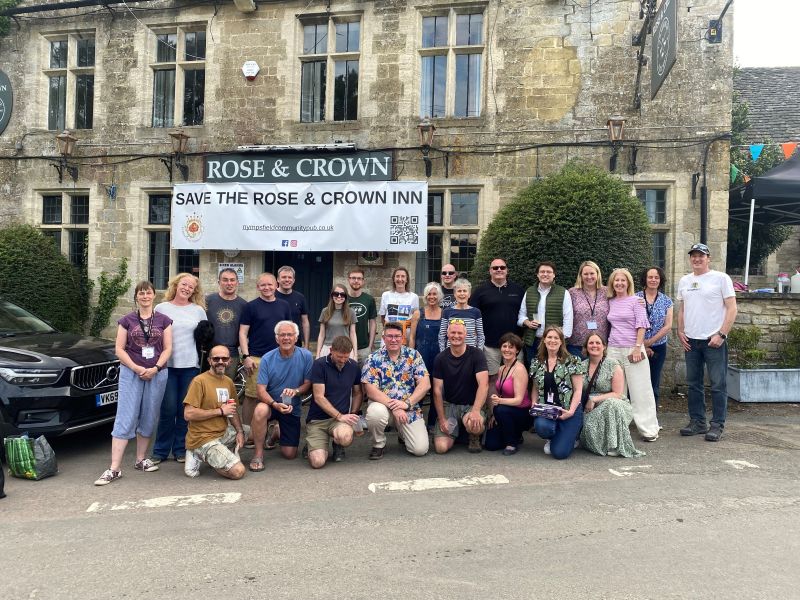 Picture showing a number of people looking ahead. There is an old rustic building behind them with a banner that says 'ROSE & CROWN'