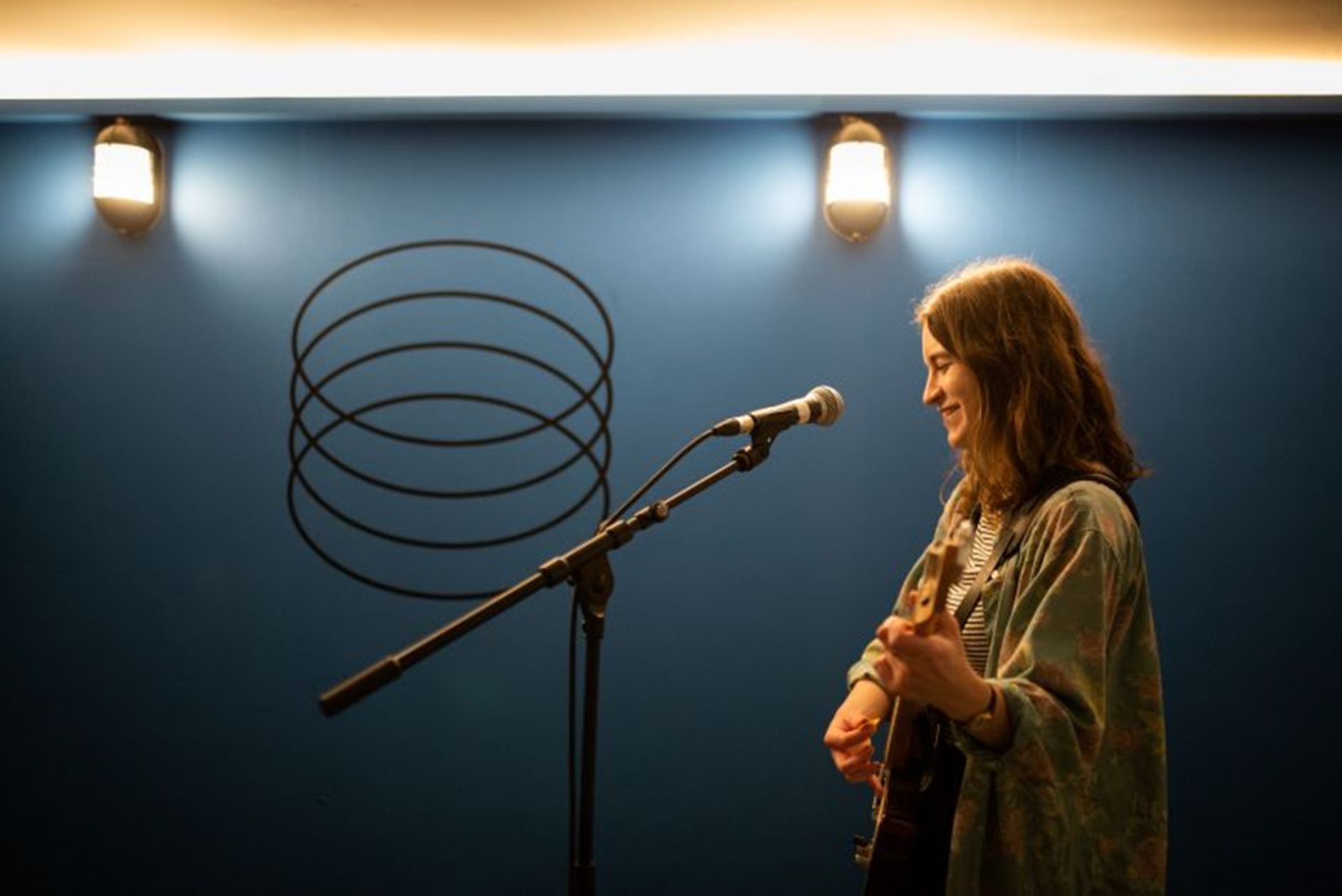 Woman smiling while playing the guitar and singing. A mic placed in front of her. She's in a room with dark blue walls