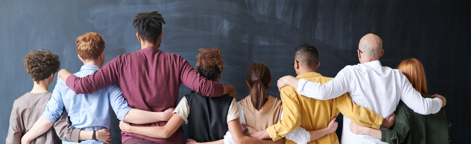 A row of people standing in a line facing a blackboard with their arms around each other
