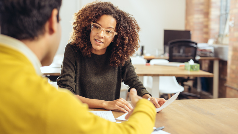 Woman looks at a sheet of paper while sat at a desk with the back of a person facing her