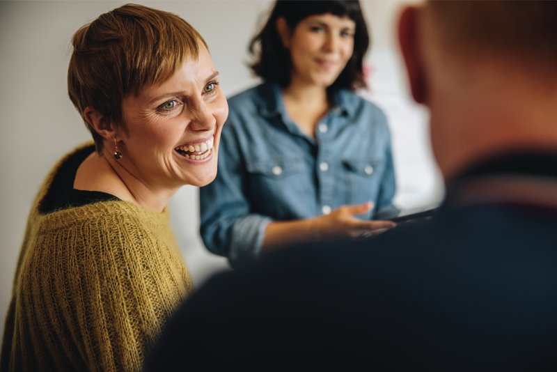 The image shows a woman with short, light brown hair wearing a mustard-coloured jumper, smiling as she engages in conversation. In the background, another woman with dark hair, dressed in a denim shirt, looks on with a gentle expression. The scene appears casual and friendly, with a relaxed atmosphere.