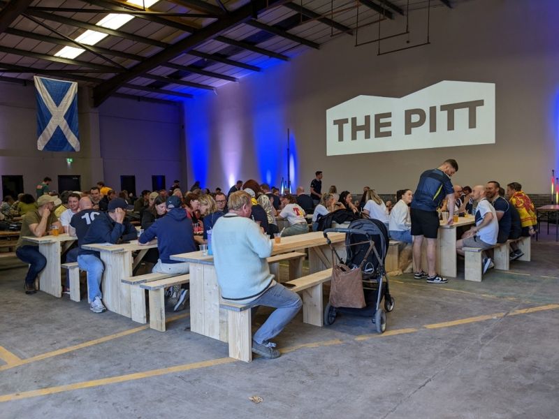 A large indoor food hall with wooden picnic-style benches filled with people eating and socializing. A projection on the wall reads "THE PITT" in bold letters. The space has industrial ceilings and is lit with blue and purple uplighting. A stroller is parked near one of the tables, and a large Scottish flag hangs on the back wall.
