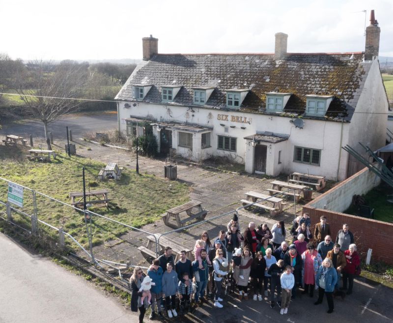 The image shows a gathering of people and a rustic white house behind them, next to them a garden.