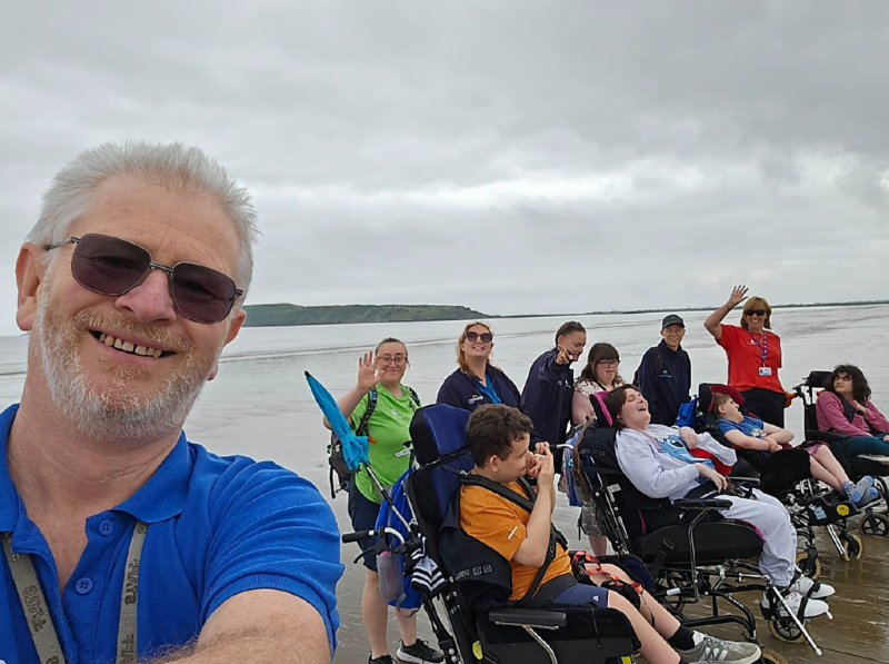 Man to the left of the image taking a selfie with a group of people at the seaside