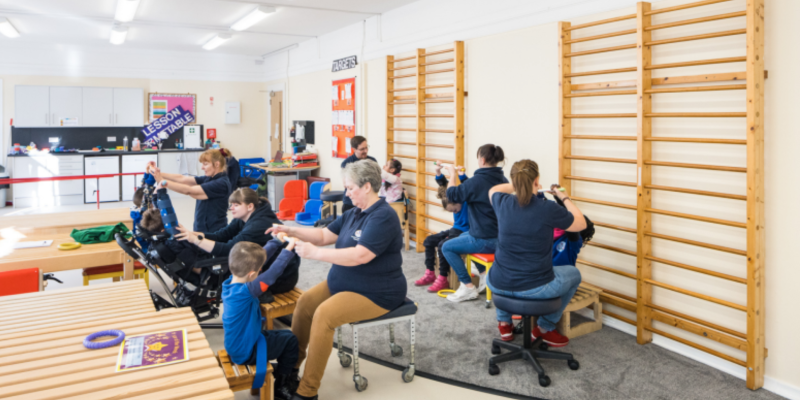 A group of adults and children in a brightly lit classroom setting. The adults are assisting the children with various activities, some of whom are using adaptive equipment. Wooden climbing bars are mounted on the wall, and there are benches, tables, and colourful educational tools in the background.