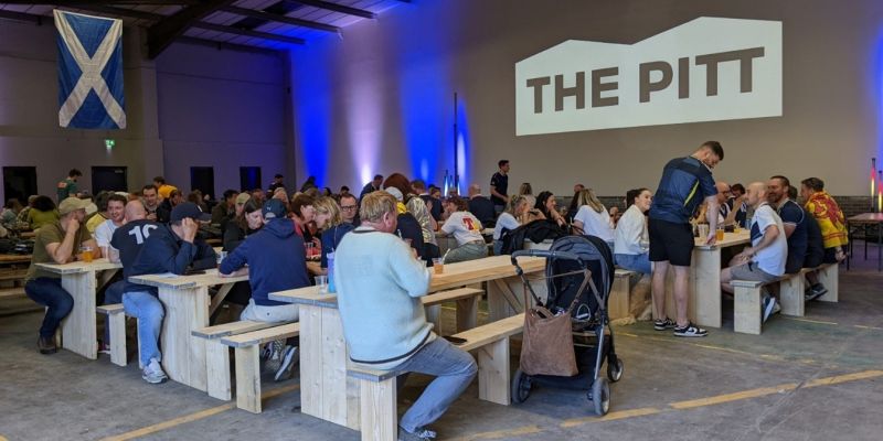 A large indoor food hall with wooden picnic-style benches filled with people eating and socializing. A projection on the wall reads "THE PITT" in bold letters. The space has industrial ceilings and is lit with blue and purple uplighting. A stroller is parked near one of the tables, and a large Scottish flag hangs on the back wall.