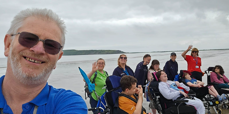 Man to the left of the image taking a selfie with a group of people at the seaside