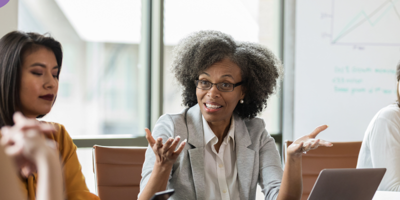 A group of women at a table in conversation. One woman is gesturing as if to ask a question.