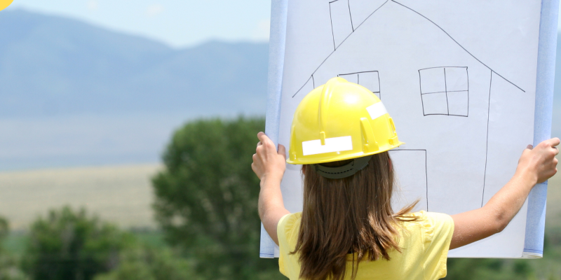 A little girl wearing a hard hat and high vis jacket looks at a drawing of a house